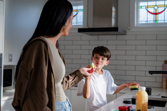 Maori mum handing a slice of apple to young son washing dishes in the kitchen