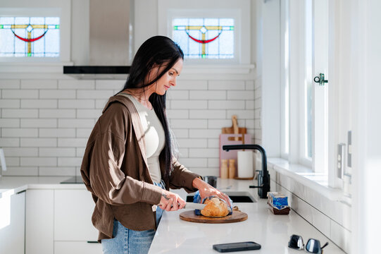 A woman stands in a kitchen cutting bread on a wooden board while sunlight enters through windows