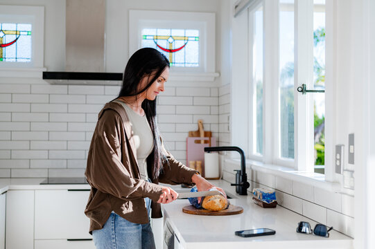 A woman stands by a kitchen counter cutting bread while sunlight streams in through the windows