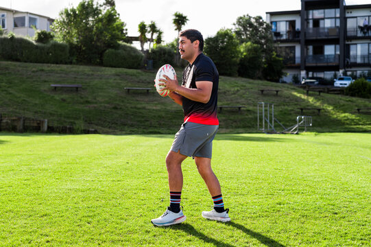 Samoan man stands on green grass, getting ready to throw a rugby ball in a sunny park