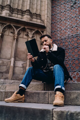 Fototapeta premium man working on tablet sitting on stairs, male student studying online in historic city, digital nomad, 5G internet technology, wireless connection, lifestyle