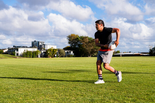 Samoan man running and throwing a rugby ball on a grassy field under a clear sky