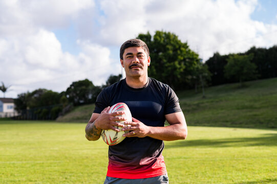 Samoan rugby player stands with a ball on a green field during a sunny day, smiling