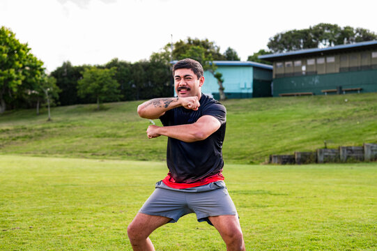 A man engages in a cultural dance on a green field surrounded by trees and buildings
