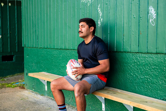 Samoan man sitting on wooden bench holding a rugby ball in waiting area