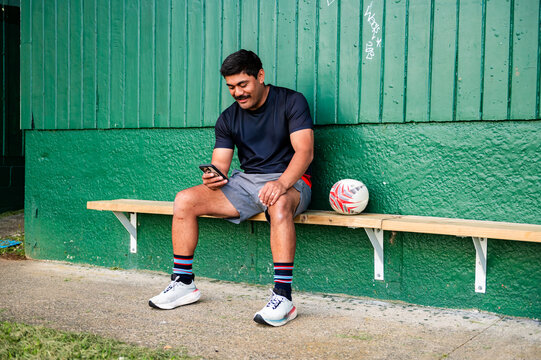 Samoan rugby player sitting on wooden bench holding a phone