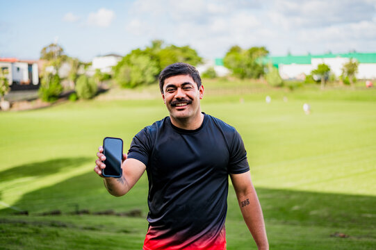 Smiling man in sportswear shows off his phone in a sunny outdoor setting with grass