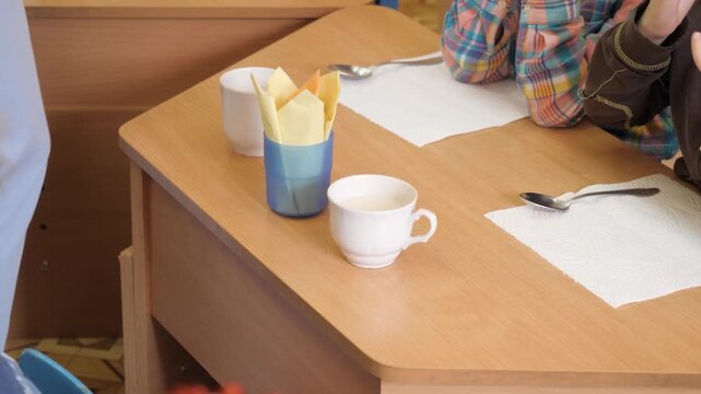 Adult hand pouring hot white milk beverage from vintage beige enamel teapot into ceramic cup for preschool students sitting at wooden table during morning breakfast snack time