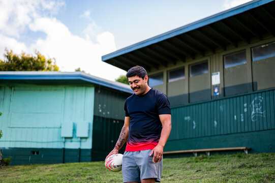 A man stands smiling while holding a footy, enjoying his time at a sports field near a building