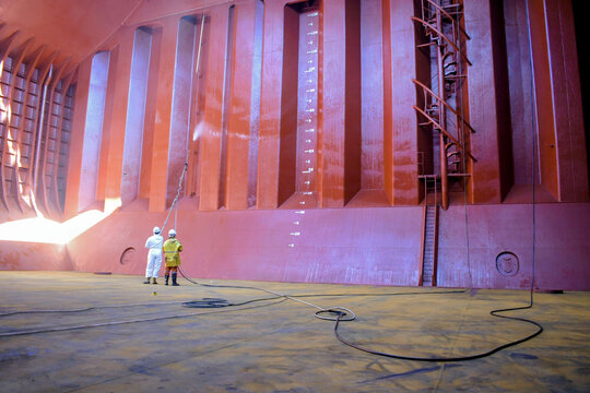 crew members in protective suits spray chemical coating on cargo hold bulkhead for corrosion protection and maintenance