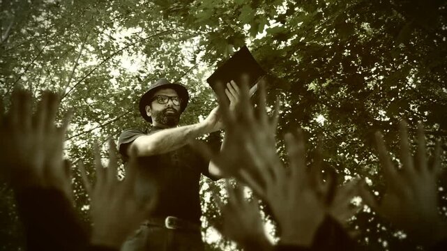 Religious man, reverend or priest wearing a clerical collar and thumping a bible to his congregation. Preacher preaching the gospel to a group of followers in a rural setting.