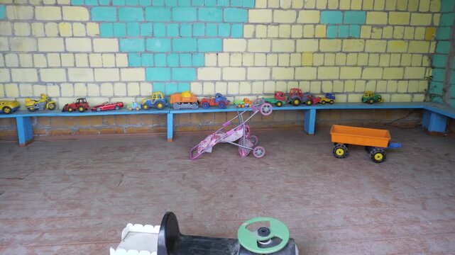 Row of vibrant plastic toy cars trucks and strollers organized on blue wooden shelf inside outdoor kindergarten pavilion during sunny summer day.