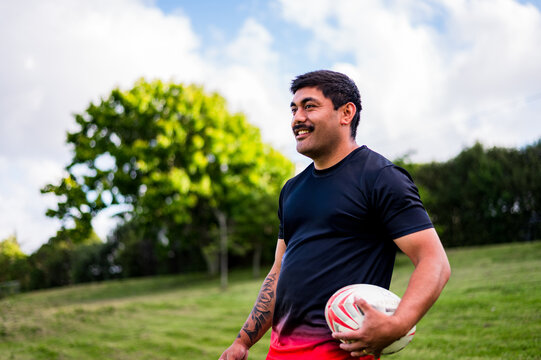 A male rugby player stands on a grassy field, smiling as he holds a rugby ball