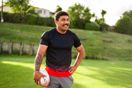A rugby player smiles while standing on a grassy field holding a ball