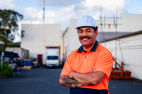 A cheerful construction worker poses with crossed arms at a busy building site in Auckland