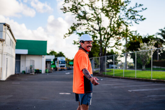 Worker in orange shirt and helmet smiles while standing in a busy outdoor work site