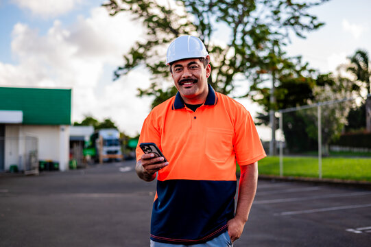 A worker stands confidently, looking at his phone in vibrant safety gear at a job site