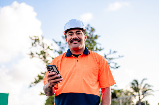 Man in high-visibility clothing holds phone, enjoying a sunny day outdoors at a work site