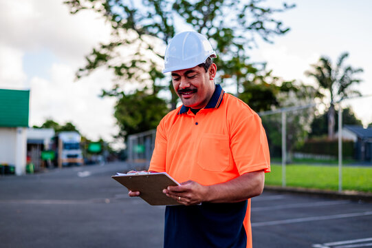 A construction worker checks plans while standing in a yard, wearing safety gear and smiling