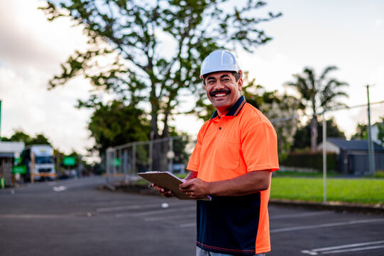 A young construction manager holds a clipboard and smiles at a worksite during sunset