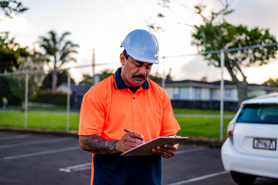 A construction worker in high-visibility gear is writing on a clipboard in a car park