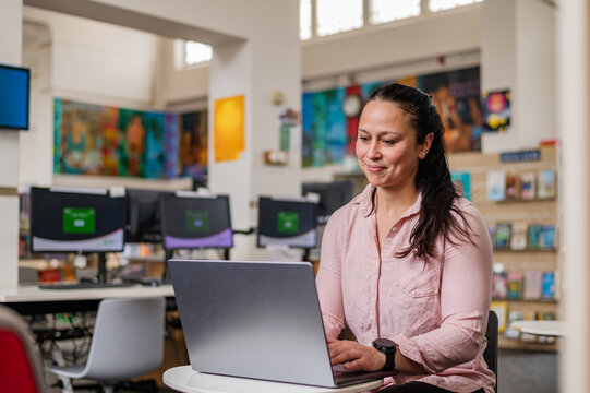 Woman sits at table in library, focused on laptop while studying and smiling