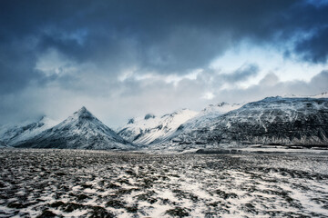 A wide-angle landscape featuring jagged snow-covered mountains and a frozen tundra under a heavy, dramatic winter sky. Ideal for travel and nature themes. © marcodenaro