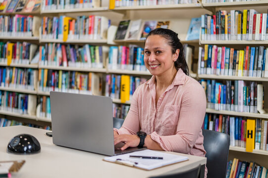 A woman sits at a desk in a library typing on a laptop while smiling