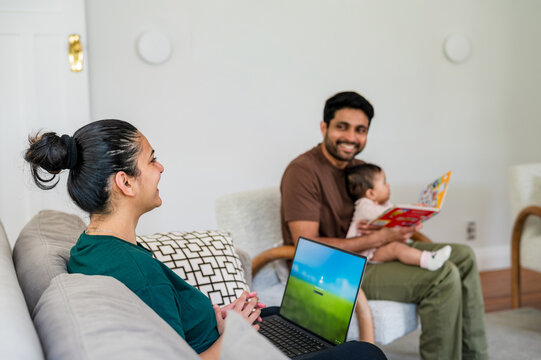 Indian mum working on her laptop while dad reads a book to daughter in the living room
