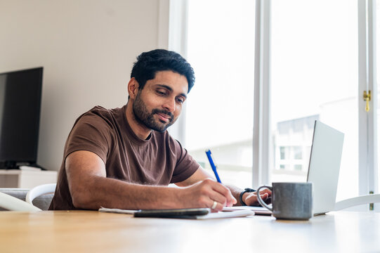 Indian man writing notes while looking at a laptop on the table