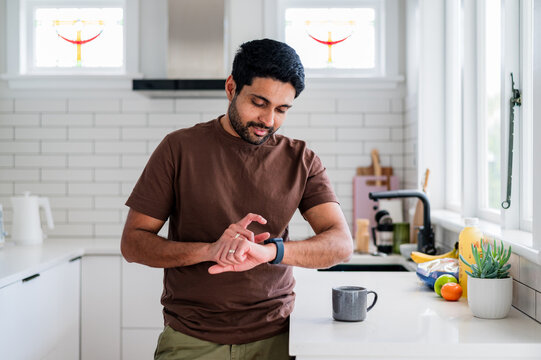 Indian man standing beside the kitchen counter looking and tapping on his smartwatch