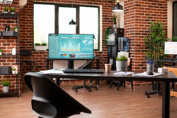 Fototapeta premium Empty startup office having modern desk with computer monitor displaying financial charts. Desktop pc, shelves with folders, boxes and plant decorations completing well organized brick wall workspace.