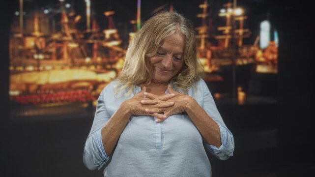 Senior woman clasping hands over chest, smiling with closed eyes in a studio with a lit ship exhibit and warm soft lighting; contentment.