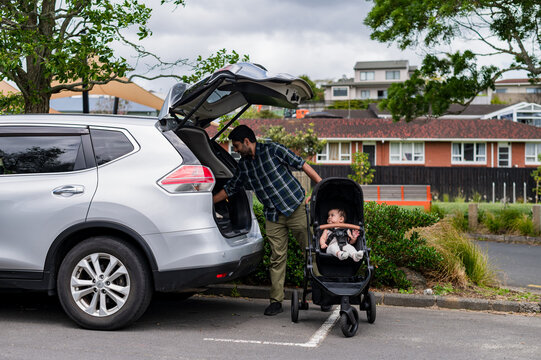 Child waits in stroller as father is fixing items into the back of a car at a park setting