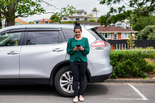 A South Asian woman in casual clothing is standing near a parked SUV, focused on her mobile phone