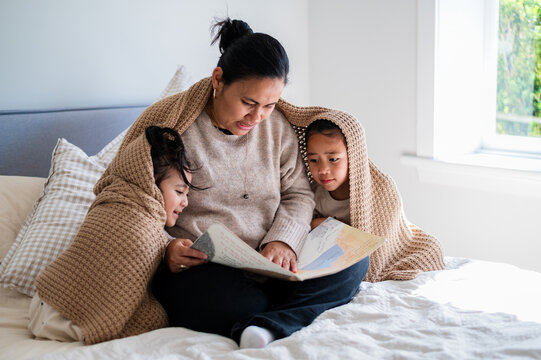 Niuean mum with her young daughters sitting closely on the bed wrapped in a blanket reading a book