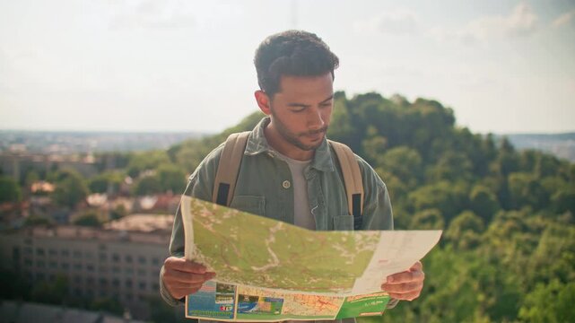 Young Caucasian lost male tourist with backpack holding reading paper map on hilltop. Bearded man in green shirt studying route. Panoramic city and forest background. Outdoors. Travel concept.