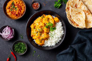 Traditional Indian meal: chicken tikka masala over rice, naan bread, lentil curry, fresh garnishes. Dark background. Close up. Top view. © annapustynnikova
