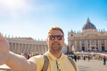 Smiling male tourist taking selfie in St Peter Square with St Peter Basilica in background on sunny...