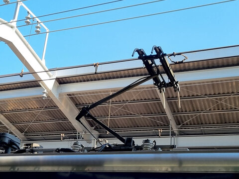 Close up of an electric train pantograph connected to overhead power lines at a railway station. Industrial details of public transportation infrastructure against a clear blue sky background.