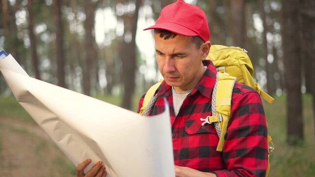 Reading map in forest with backpack man wearing red cap and plaid shirt studies route during hike on pine trail navigation task for traveler exploring woodland path map study navigation planning
