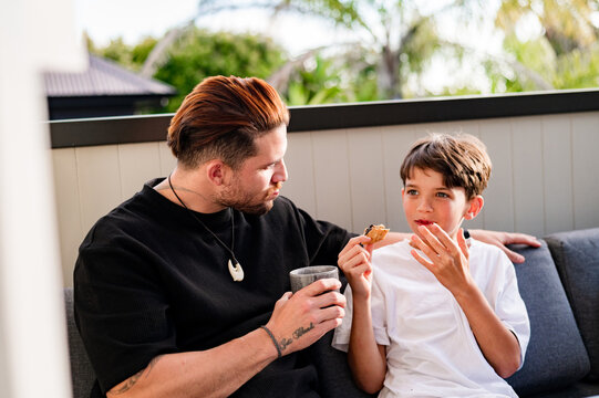 Father sharing a meal with tween son while seated on the couch in the balcony