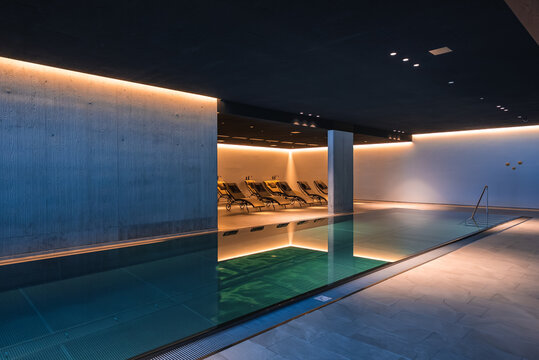 Sleek indoor spa pool glows with warm indirect lighting, smooth concrete walls, and a row of cushioned lounge chairs in a luxury Dolomites hotel at evening light.