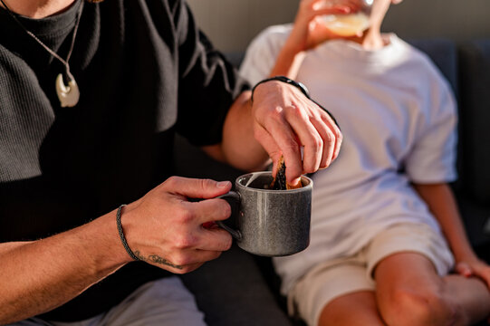 Adult hand dipping a biscuit in a cup while a young boy sits nearby drinking.