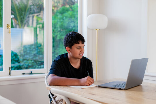 Teenage boy doing his homework on wooden table writing behind open laptop