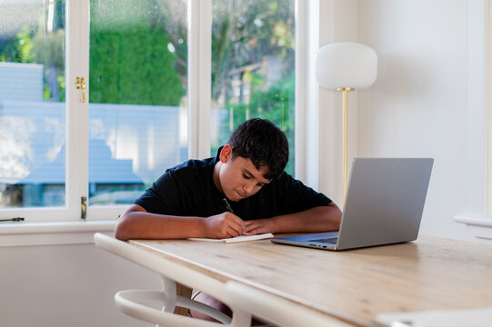 Teenage boy doing his homework on wooden table writing behind open laptop