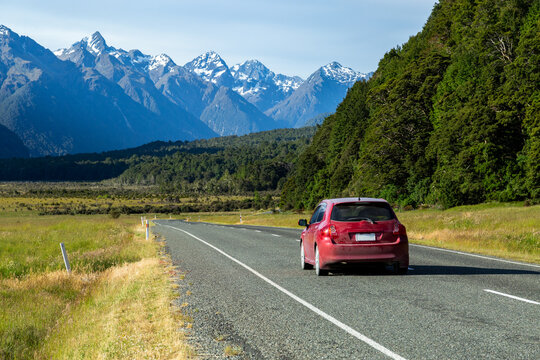 Red compact car driving down road with snow-capped mountains in background