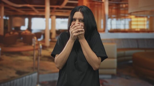 Woman covering mouth with hands laughing in airport lounge, shoulders raised and eyes squinting; amusement surprise.
