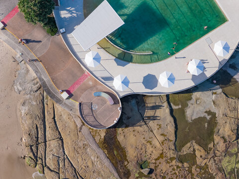 Aerial view of people swimming in a coastal pool with umbrellas alongside and a raised lookout