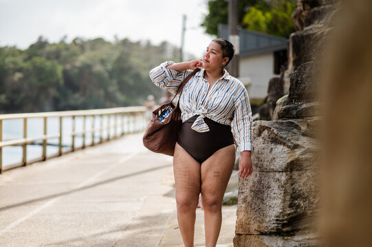 Curvy woman walking on coastal path carrying a brown beach bag on sunny day